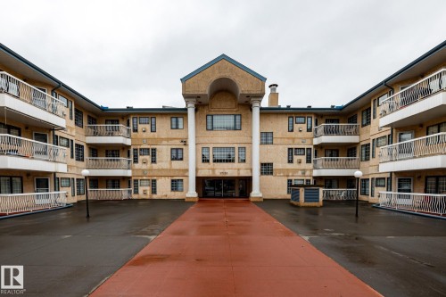 Multi-story building featuring numerous balconies with white railings, a central entrance with large columns, and a paved courtyard with a distinct red pathway - #310 17151 94A Av Nw, Edmonton, AB - Outdoor With Balcony With Facade