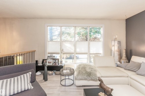 Living area featuring large multi-panel windows with cellular shades, light-toned walls with one accent wall, light wood-finish flooring, and a contemporary railing with wood top and black balusters - 13208 128 Avenue Nw, Edmonton, AB - Indoor Photo Showing Living Room