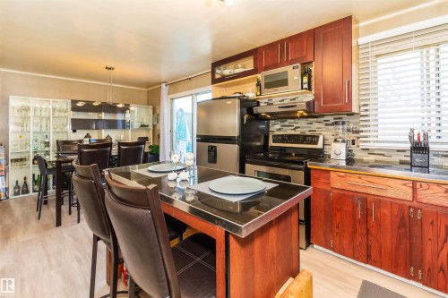 Kitchen featuring a central island with a dark countertop, stainless steel appliances, upper cabinetry, lower wood-finish cabinetry, and a tile backsplash - 13208 128 Avenue Nw, Edmonton, AB - Indoor