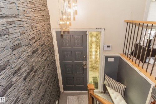 Entryway featuring a stacked stone accent wall, a modern linear chandelier, a panel door with sidelight, light wood flooring, and a contemporary staircase with metal balusters - 13208 128 Avenue Nw, Edmonton, AB - Indoor Photo Showing Other Room