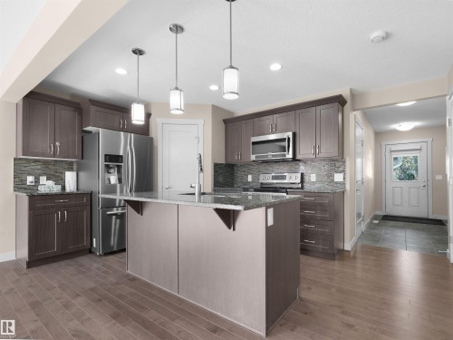 Contemporary kitchen featuring a central island with a sink, stainless steel appliances, dark wood-finish cabinetry, stone countertops, and wood-finish flooring - 4487 Annett Common Sw, Edmonton, AB - Indoor Photo Showing Kitchen With Stainless Steel Kitchen With Upgraded Kitchen