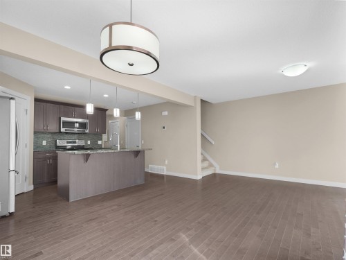 Open-concept living area featuring wood-finish flooring, a contemporary drum pendant light, and a kitchen with stainless steel appliances, dark wood cabinetry, and a central island with pendant lighting - 4487 Annett Common Sw, Edmonton, AB - Indoor Photo Showing Kitchen