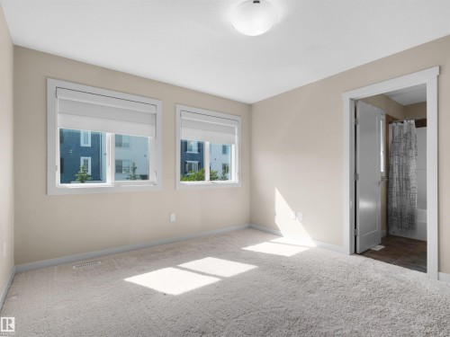 Carpeted room featuring two windows with roller blinds, a ceiling-mounted light fixture, and a doorway revealing a partial bathroom with a shower-tub combination and tiled flooring - 4487 Annett Common Sw, Edmonton, AB - Indoor