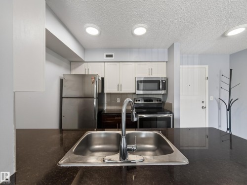 Kitchen featuring stainless steel appliances, light cabinetry, and dark countertops - #217 12045 22 Av Sw, Edmonton, AB - Indoor Photo Showing Kitchen With Stainless Steel Kitchen With Double Sink