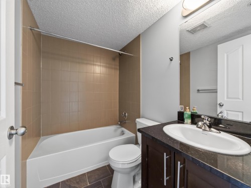 Bathroom featuring a white tub, shower with tan tile surround, white toilet, dark wood-finish vanity with a black countertop, and a chrome faucet - #217 12045 22 Av Sw, Edmonton, AB - Indoor Photo Showing Bathroom