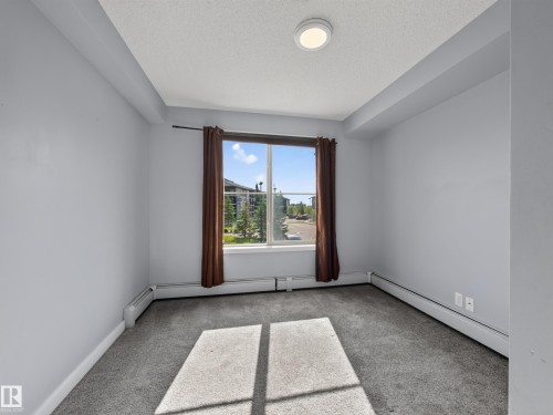 Carpeted room featuring a large window, neutral wall paint, and baseboard heating - #217 12045 22 Av Sw, Edmonton, AB - Indoor Photo Showing Other Room