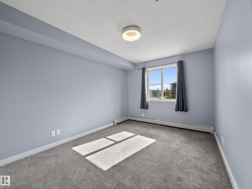Room with light-toned walls, grey carpeting, and a window with a building and greenery visible - #217 12045 22 Av Sw, Edmonton, AB - Indoor Photo Showing Other Room