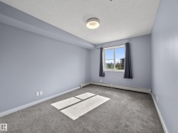 Room with light-toned walls, grey carpeting, and a window with a building and greenery visible - 