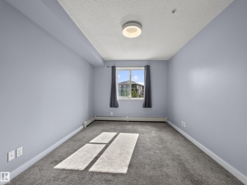 Spacious room featuring light blue walls, gray carpet, and a window offering exterior views - #217 12045 22 Av Sw, Edmonton, AB - Indoor Photo Showing Other Room