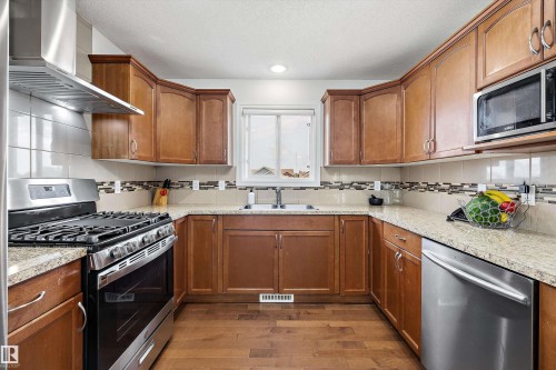 U-shaped kitchen featuring wood-finish cabinetry, light granite countertops, and stainless steel appliances including a gas range with an overhead hood fan, and a built-in dishwasher - 2803 12 Street Nw, Edmonton, AB - Indoor Photo Showing Kitchen With Double Sink