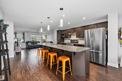 Open-concept kitchen featuring an oversized island with pendant lighting, granite countertops, wood-finish flooring, dark wood cabinetry, and stainless steel appliances - 