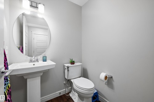 Pedestal sink with chrome fixtures and an oval mirror, complemented by wood-finish flooring, a white toilet, and light gray wall paint - 1563 Tamarack Boulevard Nw, Edmonton, AB - Indoor Photo Showing Bathroom