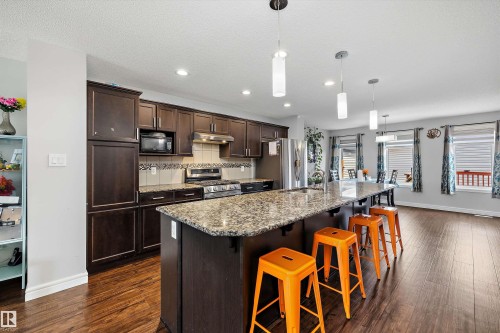 Open-concept kitchen featuring wood-finish flooring, dark wood cabinetry, granite countertops, a tiled backsplash, and stainless steel appliances - 1563 Tamarack Boulevard Nw, Edmonton, AB - Indoor Photo Showing Kitchen With Stainless Steel Kitchen With Upgraded Kitchen
