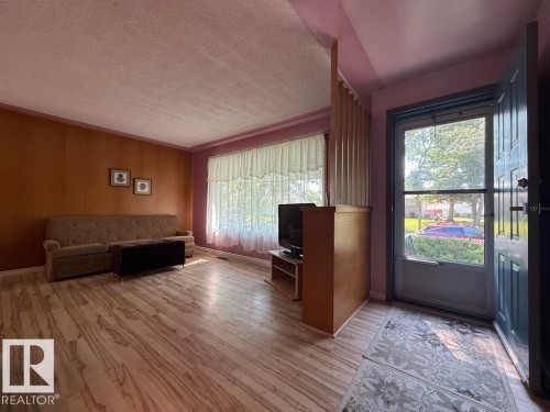Spacious living area featuring wood-finish flooring, a full wall of wood paneling, and a large front window with sheer curtains - 9908 96A Avenue, Fort Saskatchewan, AB - Indoor