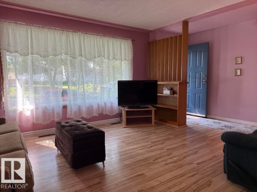 Spacious living area featuring wood-finish flooring, a large window with white lace curtains, and a built-in room divider with shelving - 9908 96A Avenue, Fort Saskatchewan, AB - Indoor Photo Showing Living Room