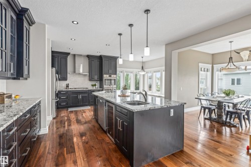 Gourmet kitchen featuring dark wood cabinetry, granite countertops, an expansive center island with a built-in beverage cooler, stainless steel appliances, and wood-finish flooring - 1421 Westerra Bay, Stony Plain, AB - Indoor Photo Showing Kitchen With Upgraded Kitchen