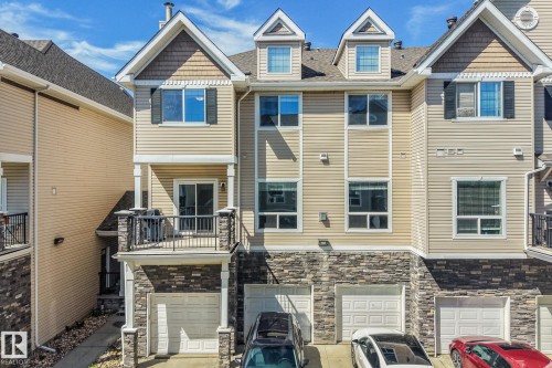 Multi-level townhouse exterior featuring stacked stone accents, neutral siding, a private balcony with black railings, a second-story sliding glass door, and a ground-level attached garage - #16 7293 South Terwillegar Dr Nw, Edmonton, AB - Outdoor With Facade