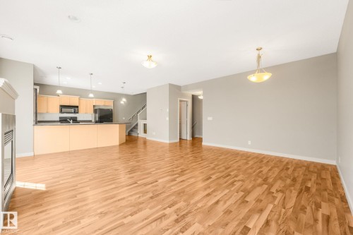 Open-concept living area with wood-finish flooring, a contemporary fireplace, and a kitchen featuring light wood cabinetry, dark countertops, and black appliances - #16 7293 South Terwillegar Dr Nw, Edmonton, AB - Indoor Photo Showing Kitchen