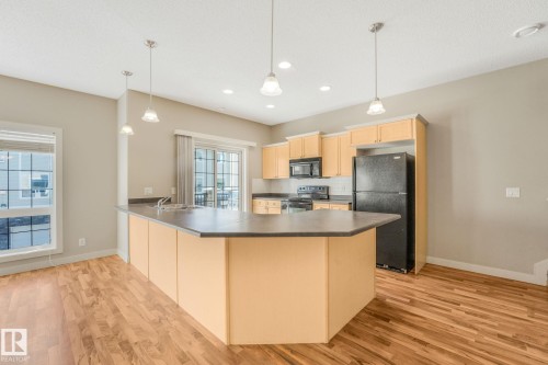 Kitchen featuring wood-finish flooring, a central island with a dark countertop, light wood cabinetry, black appliances, and pendant lighting - #16 7293 South Terwillegar Dr Nw, Edmonton, AB - Indoor Photo Showing Kitchen