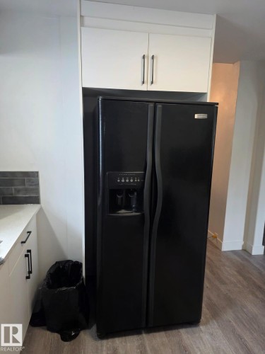 Black side-by-side refrigerator with an ice and water dispenser, integrated into a kitchen featuring white upper cabinetry with dark hardware and wood-finish flooring - 10656 93 Street, Edmonton, AB 
