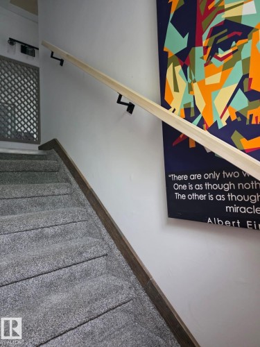Carpeted staircase featuring a light wood-finish handrail with dark metal brackets, white painted walls, and wood-finish trim along the stair stringer - 10656 93 Street, Edmonton, AB 