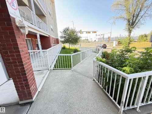 Exterior entrance featuring a concrete ramp with white metal railings, brick column accents, and surrounding greenery - #302 8215 84 Av Nw, Edmonton, AB - Outdoor With Balcony With Exterior