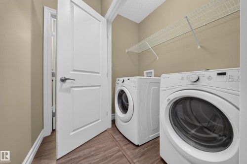 Dedicated laundry space featuring a front-load washer and dryer, wood-finish flooring, neutral-toned walls, and a wire storage shelf - #100 804 Welsh Dr Sw, Edmonton, AB - Indoor Photo Showing Laundry Room