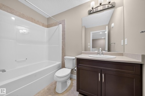 Bathroom featuring a white tub-shower combination with a tiled surround, a toilet, a dark wood-finish vanity with a light countertop, an undermount sink, and a large wall-mounted mirror - #100 804 Welsh Dr Sw, Edmonton, AB - Indoor Photo Showing Bathroom
