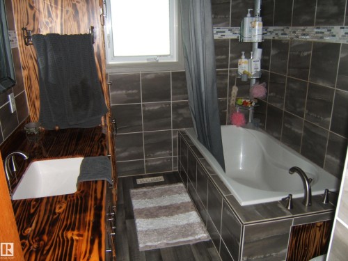 Bathroom featuring a soaking tub with dark tile surround, a built-in wood-finish vanity with an integrated sink, dark wood-finish flooring, and a tiled accent wall - 151042 Twp  Rd 562, Rural Two Hills County, AB - Indoor Photo Showing Bathroom