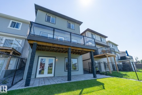 Exterior rear elevation featuring a spacious upper-level deck with glass panel railings, a lower-level paver patio, double glass-paneled doors, and a manicured lawn - 5403 Lark Landing Nw, Edmonton, AB - Outdoor With Deck Patio Veranda