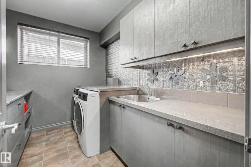 Dedicated laundry area featuring a stainless steel sink, ample gray cabinetry, light-toned countertops, and a decorative textured backsplash - 38 Longview Point(E), Spruce Grove, AB - Indoor Photo Showing Laundry Room
