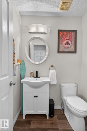 Bathroom featuring a white vanity with integrated sink, a matching white toilet, a round wall-mounted mirror, and wood-finish flooring - 833 Mcallister Crescent Sw, Edmonton, AB - Indoor Photo Showing Bathroom