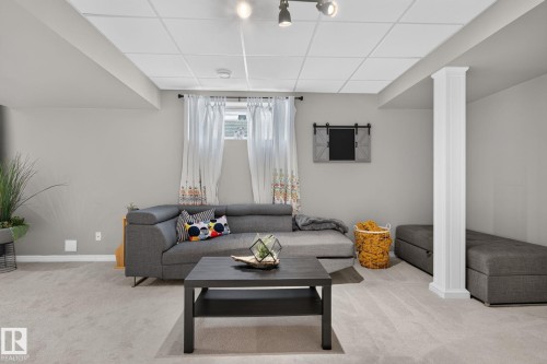 Carpeted living space featuring a drop ceiling with track lighting, a window with white curtains, and a decorative fluted column - 833 Mcallister Crescent Sw, Edmonton, AB - Indoor Photo Showing Living Room