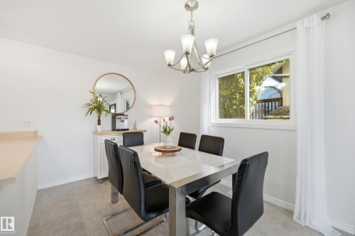 Bright dining area featuring a modern chandelier, a large window with white sheer curtains, light wall-to-wall carpeting, and a built-in countertop extension - 15111 67 Street Nw, Edmonton, AB - Indoor Photo Showing Dining Room
