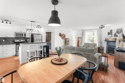 Open-concept living space featuring wood-finish flooring, a kitchen with white cabinetry and stainless steel appliances, a breakfast bar, a dining area, and a living room with a fireplace - 679 Kananaskis Drive, Devon, AB - Indoor With Fireplace