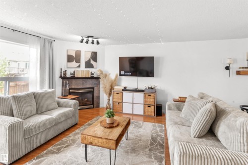 Living area featuring wood-finish flooring, a fireplace with a dark stone surround and mantel, track lighting, and a large window with white sheer curtains - 679 Kananaskis Drive, Devon, AB - Indoor Photo Showing Living Room With Fireplace