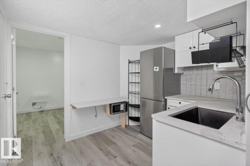 Compact kitchen area featuring wood-finish flooring, white cabinetry, a stainless steel refrigerator, and a dark undermount sink with a gooseneck faucet - #18 345 Kirkness Rd Nw, Edmonton, AB - Indoor Photo Showing Kitchen