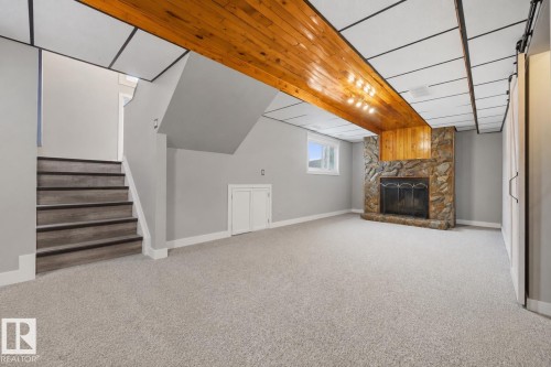 Spacious lower level featuring a stone-clad fireplace with a wood mantel, light gray carpeting, and a drop panel ceiling with a wood-finish beam - 4810 55 Avenue, Beaumont, AB - Indoor Photo Showing Other Room With Fireplace