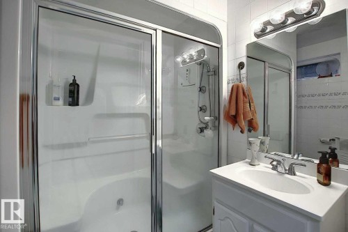 This bathroom features a shower and bathtub combination with a clear glass enclosure, a single vanity with a white countertop and basin, and a wall-mounted mirror with an overhead light fixture - 11126 52 Street Nw, Edmonton, AB - Indoor Photo Showing Bathroom