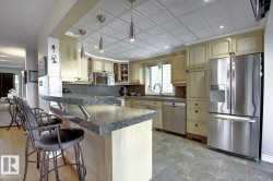 The kitchen features light wood cabinetry, a granite breakfast bar, stainless steel appliances, and a tiled ceiling with pendant lighting - 