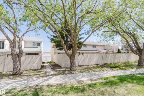 Townhouse exterior featuring light-colored siding, a white fence, and mature trees - 134 Callingwood Place Nw, Edmonton, AB - Outdoor