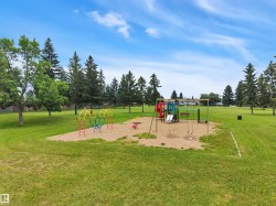 Expansive green space featuring mature trees and a dedicated playground area with slides, swings, and climbing structures on a sand base - 