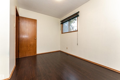 Interior room featuring dark wood-finish flooring, light-colored walls, a brown wooden door, and a single window with blinds - 4812 105B Street Nw, Edmonton, AB - Indoor Photo Showing Other Room