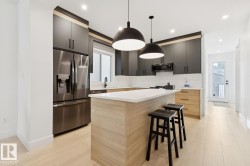Modern kitchen featuring light wood-finish flooring, dark cabinetry, stainless steel appliances, a center island with a light countertop, and dual pendant lighting - 