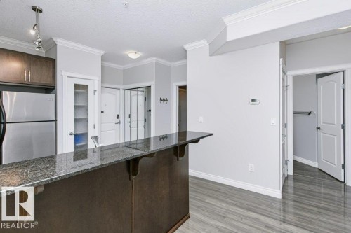 Kitchen island with a polished stone countertop, wood-finish flooring, stainless steel refrigerator, dark wood cabinetry, and crown molding - #137 10121 80 Av Nw, Edmonton, AB - Indoor Photo Showing Kitchen
