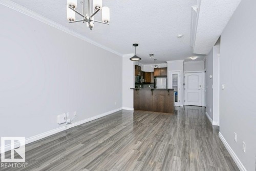 Spacious room featuring wood-finish flooring, light grey painted walls, white crown molding, and a contemporary chandelier - #137 10121 80 Av Nw, Edmonton, AB - Indoor