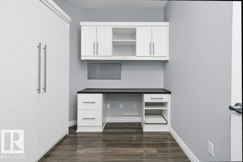 Integrated built-in desk and shelving unit with white cabinetry, dark countertop, and dark wood-finish flooring - #137 10121 80 Av Nw, Edmonton, AB - Indoor Photo Showing Other Room