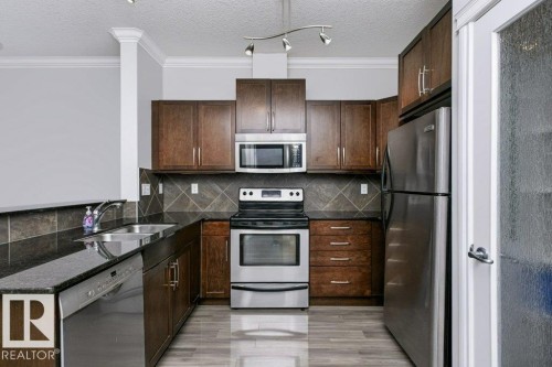 Kitchen featuring dark wood cabinetry, stainless steel appliances, dark stone countertops, and a patterned tile backsplash - #137 10121 80 Av Nw, Edmonton, AB - Indoor Photo Showing Kitchen With Stainless Steel Kitchen With Double Sink
