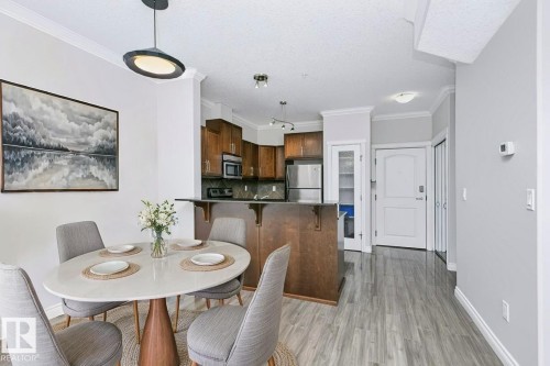 Open-concept dining and kitchen area featuring wood-finish flooring, crown molding, a kitchen island with seating, a white paneled entry door, and stainless steel appliances - #137 10121 80 Av Nw, Edmonton, AB - Indoor Photo Showing Dining Room