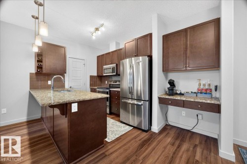 Kitchen featuring wood-finish flooring, dark wood cabinetry, a granite countertop island with pendant lighting, stainless steel appliances, and a tiled backsplash - 106 320 Ambleside Link Sw, Edmonton, AB - Indoor Photo Showing Kitchen With Upgraded Kitchen